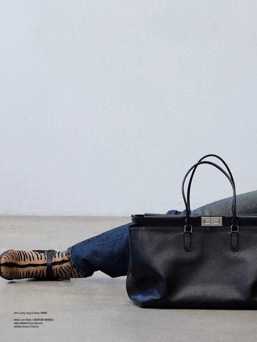 A black leather tote bag sits in the foreground with a person wearing denim jeans and tiger-striped boots reclining against a plain wall.