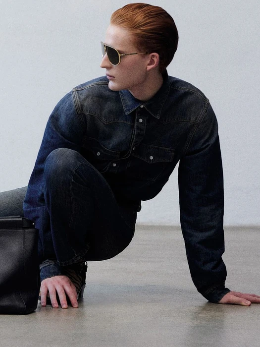 Model in dark sunglasses and denim shirt crouching next to a black handbag against a grey studio wall