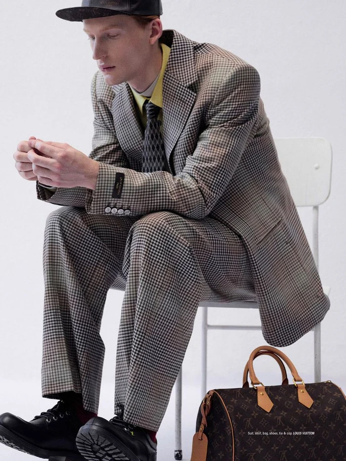 Model wearing a houndstooth suit, yellow shirt, cap and black shoes seated on a white chair next to a brown monogram duffel bag