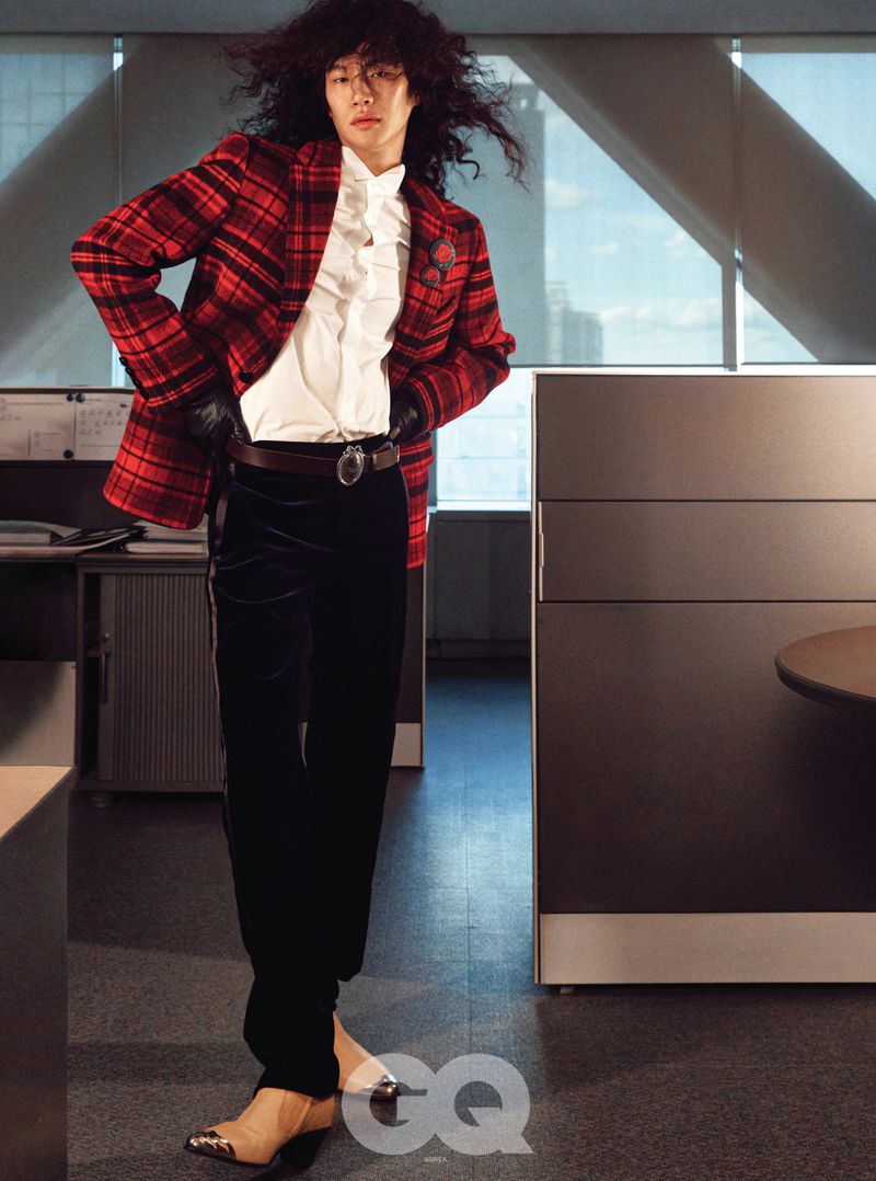 Model with long dark curly hair wearing a red plaid blazer, white shirt, and black trousers in an office setting.