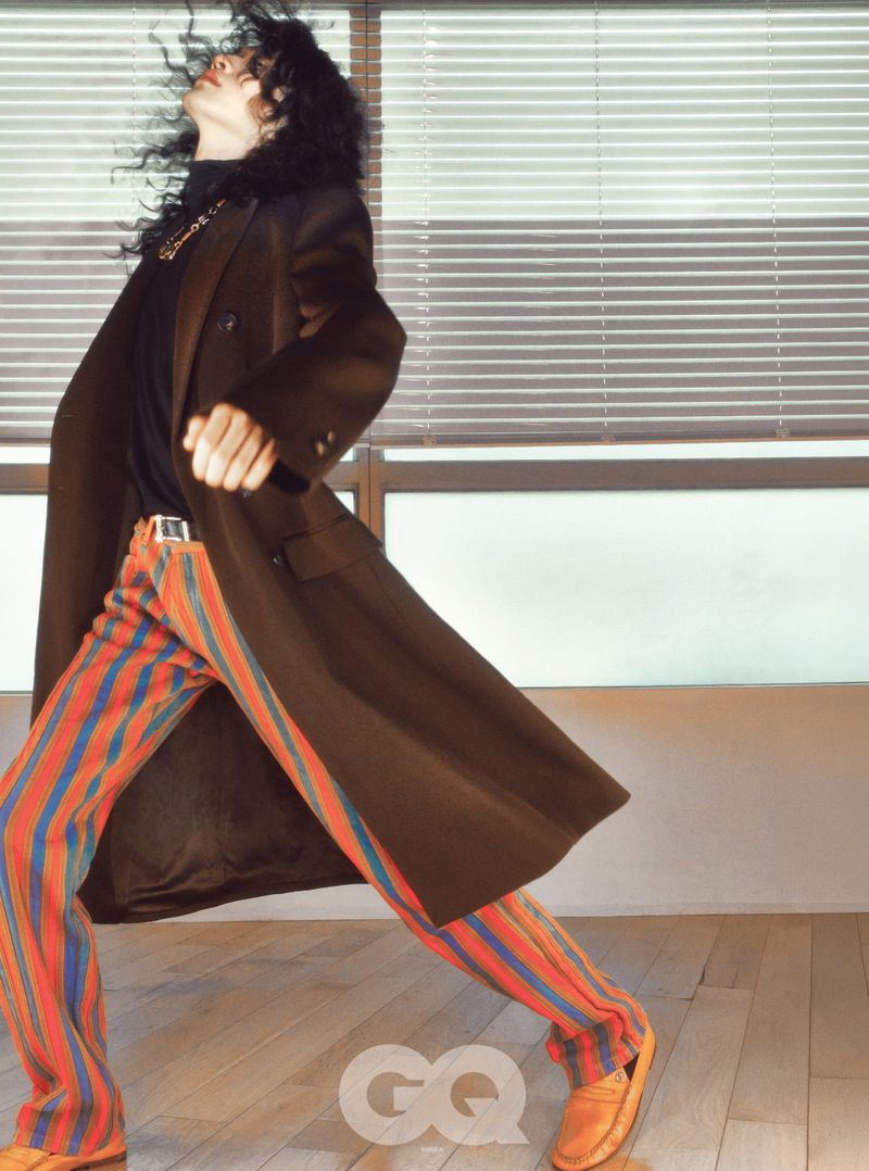 Model with dark curly hair in a brown coat and striped pants posing indoors