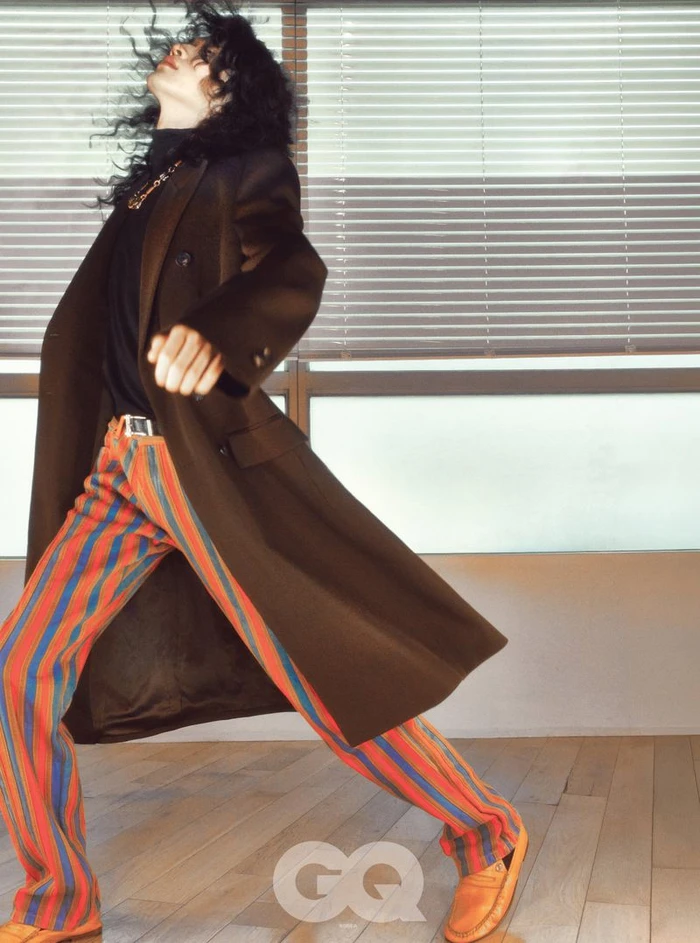 Model with dark curly hair in a brown coat and striped pants posing indoors