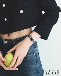 Close-up of a model wearing a black tweed jacket, denim jeans, and a luxury wristwatch while holding a green apple.