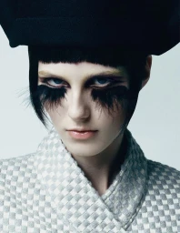 Close-up portrait of a model with heavy feathered false eyelashes, a structured black hat, and a patterned grey coat.