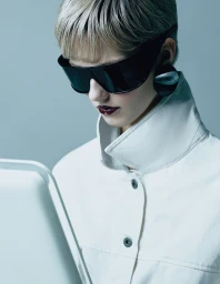 Model wearing dark sunglasses and white coat looking down in a studio setting