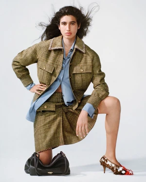 Model in olive plaid jacket and matching skirt posing with dark handbag against white seamless background