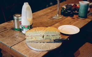 Cut Victoria sponge cake on a wooden table alongside a milk bottle and tin canister for The Gentlewoman Here project