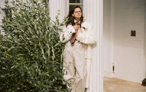 Model leaning against a white column wearing a cream coat and tie next to green foliage