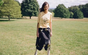 Model wearing a yellow graphic t-shirt and black skirt standing in a grassy park setting under natural sunlight.