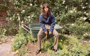 Model seated on a green wheelbarrow in a rose garden wearing a blue sweater and striped trousers