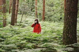 Model wearing a vibrant red coat standing amid a lush green forest of ferns, drinking from a mug.