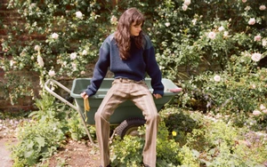Model sitting on a green wheelbarrow in a lush garden wearing a navy blue sweater and striped trousers
