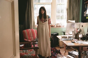 Model wearing a cream long dress standing in a home office with a computer desk and red patterned armchair