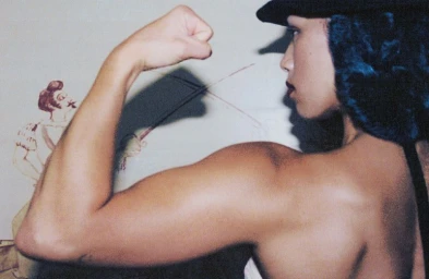 Profile view of a person flexing their bicep, wearing a dark cap and top against an illustrated wall background.