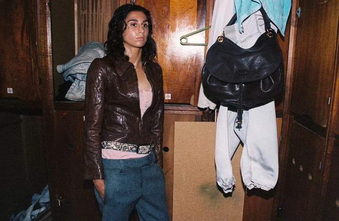 Woman in a brown leather jacket and blue trousers standing in front of wooden lockers with a black handbag hanging nearby