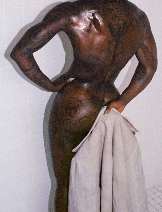 Rear view of a tattooed person holding a light-colored garment against white tiled walls