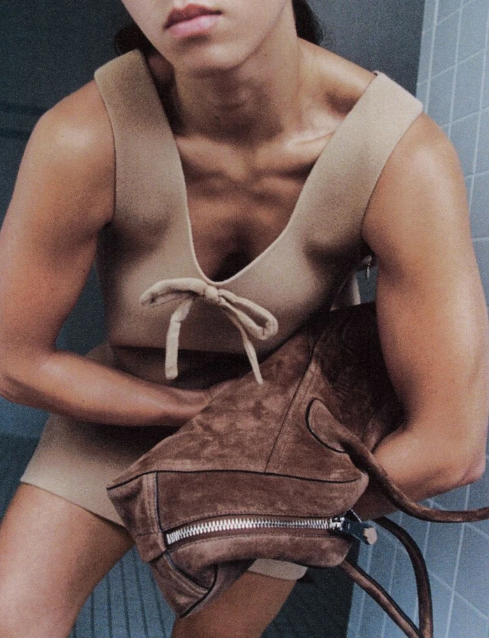 Model wearing a beige sleeveless dress and holding a brown suede handbag against a tiled wall backdrop