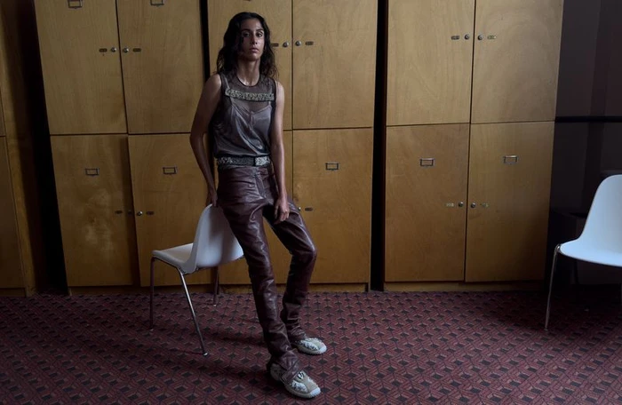 Woman leaning against a white chair in front of wooden lockers wearing leather trousers and a sheer top