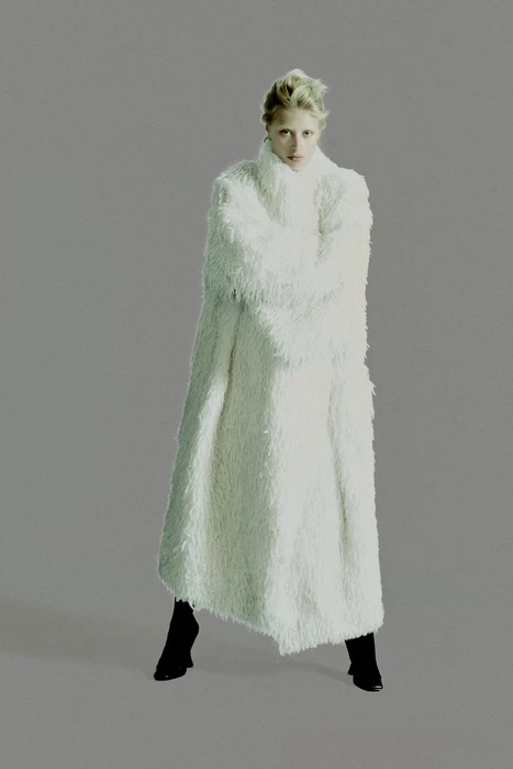 Model in a long textured white fluffy coat standing against a neutral grey studio background