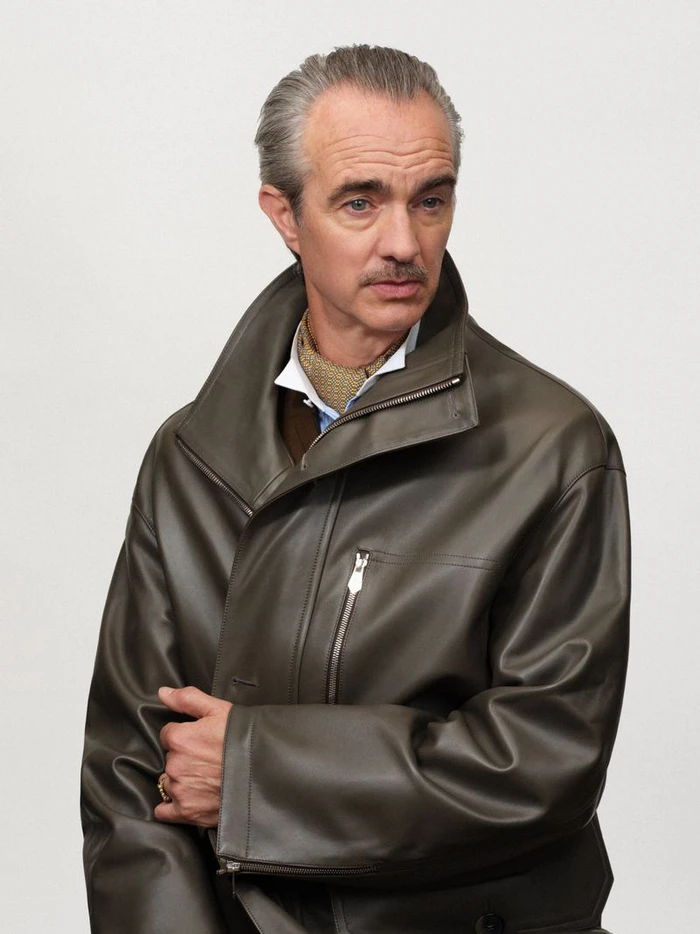 Man with slicked-back grey hair and mustache wearing an olive leather jacket over a white shirt and patterned neckerchief.