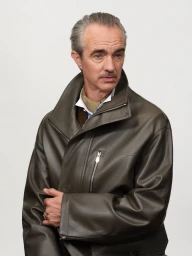 Man with slicked-back grey hair and mustache wearing an olive leather jacket over a white shirt and patterned neckerchief.