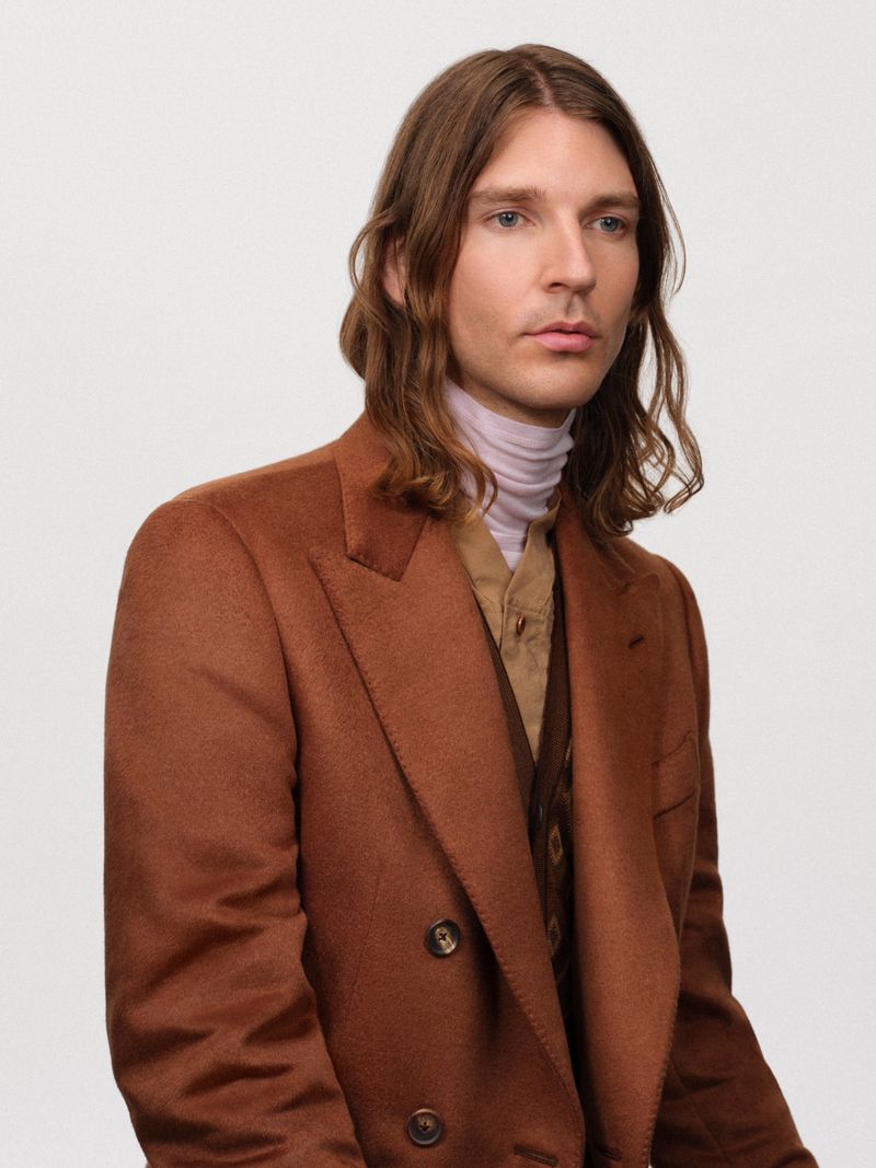 Portrait of a man with long brown hair wearing a brown blazer, tan vest, and light pink turtleneck against a white background.