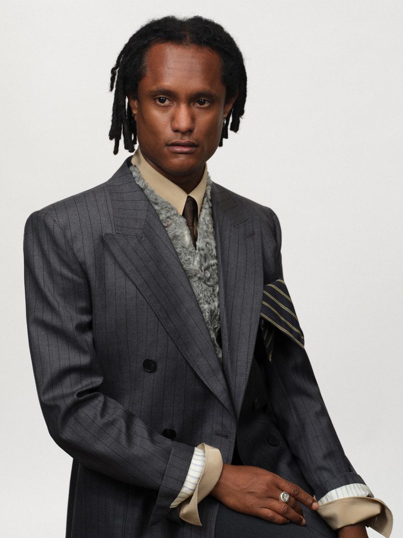 Man wearing a grey pinstripe suit jacket over a textured vest and beige shirt against a white backdrop.