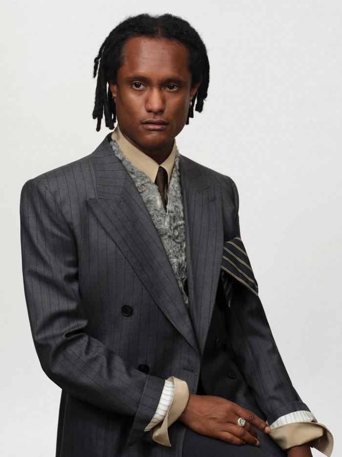 Man wearing a grey pinstripe suit jacket over a textured vest and beige shirt against a white backdrop.