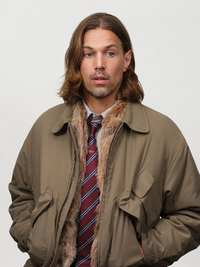 Man with long brown hair wearing an olive jacket with a fur collar and a striped tie against a plain background
