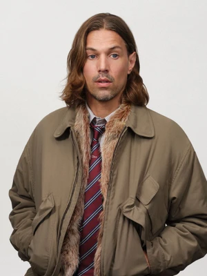 Man with long brown hair wearing an olive jacket with a fur collar and a striped tie against a plain background