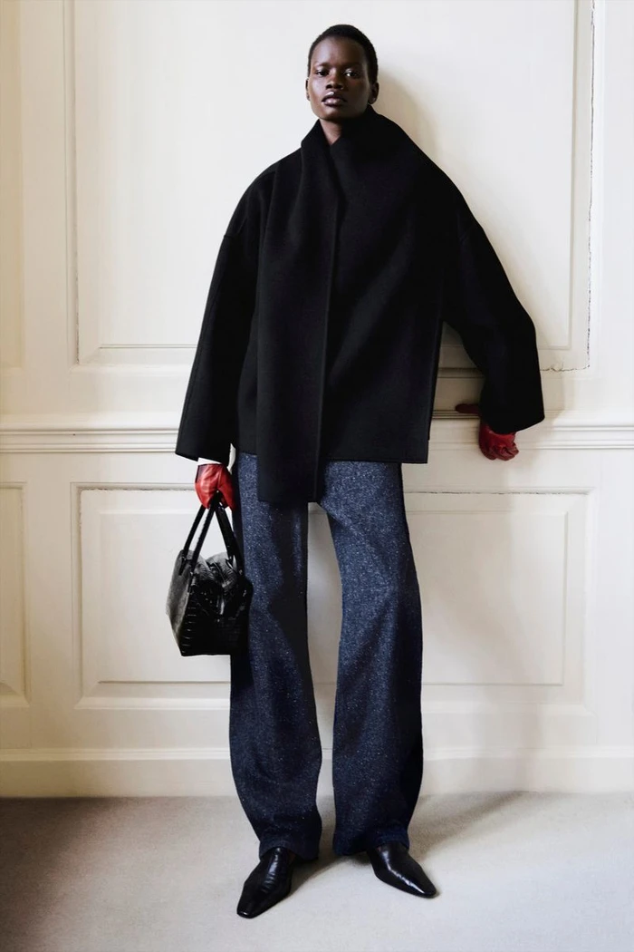 Model in black jacket and trousers holding a handbag and red gloves against an interior wall.