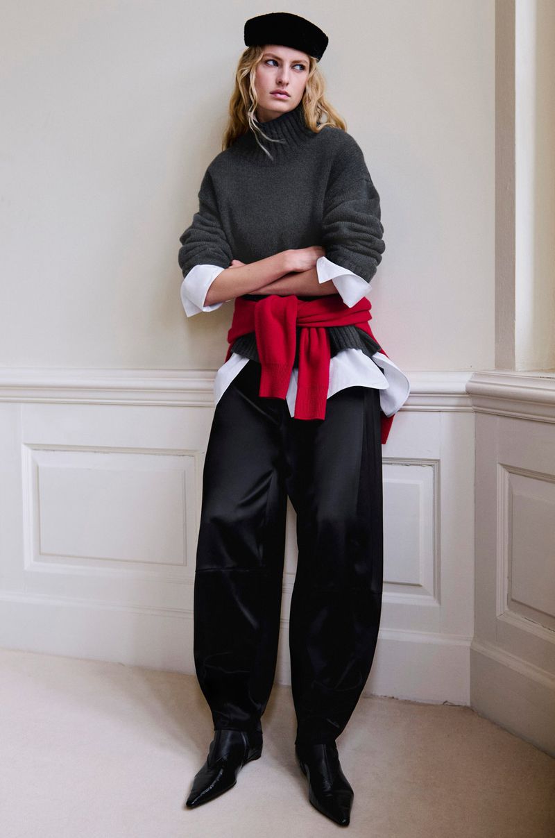 Model wearing dark grey turtleneck, white shirt, red sweater tied at waist, and black trousers against a neutral wall