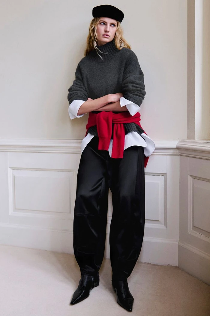 Model wearing dark grey turtleneck, white shirt, red sweater tied at waist, and black trousers against a neutral wall