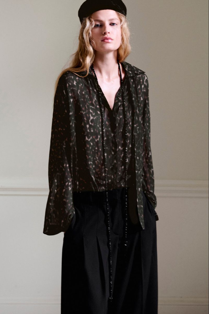 Model in patterned dark blouse and black trousers wearing a cap against a neutral interior wall