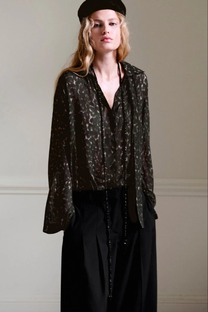 Model in patterned dark blouse and black trousers wearing a cap against a neutral interior wall