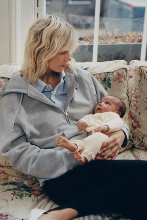 Model Marjan Jonkman in a grey zip hoodie holding a newborn baby on a floral sofa