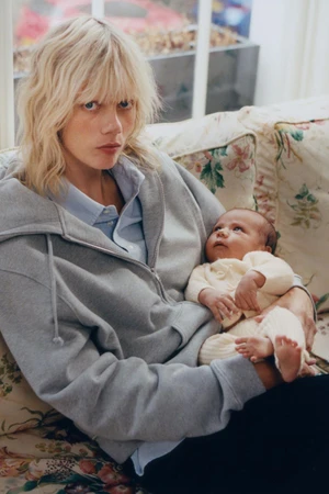 Model Marjan Jonkman wearing a grey zip hoodie holding a newborn baby on a floral sofa for Zara