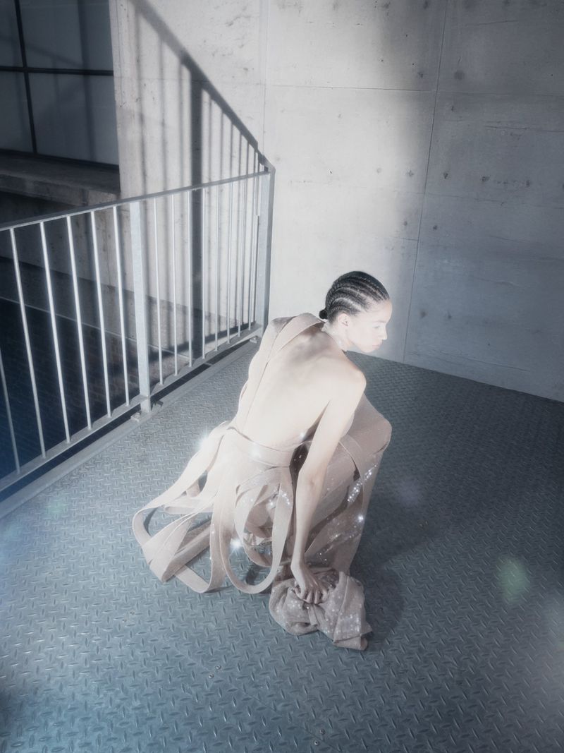 Elegant woman in beige draped fashion stands on a metal floor next to concrete walls and a metal railing, softly lit with a cool palette.