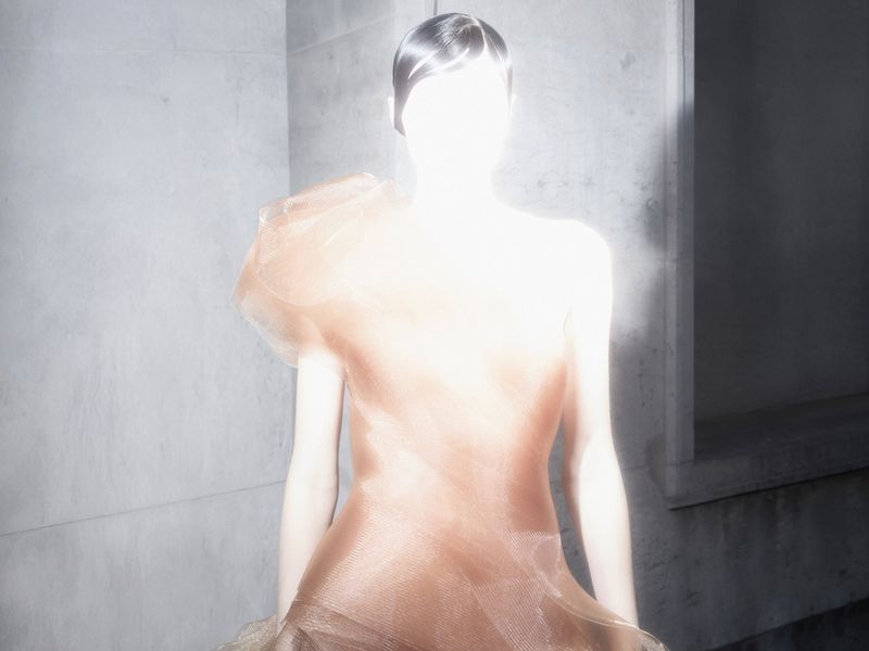 A lone figure in a textured gown stands silhouetted in a concrete corridor, the only light source behind, creating a dramatic effect.