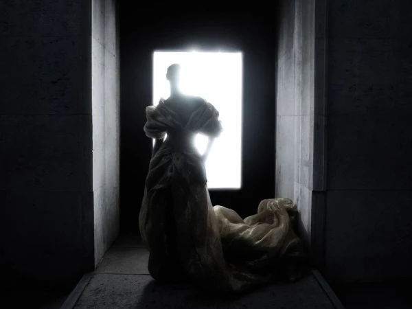 A lone figure in a textured gown stands silhouetted in a concrete corridor, the only light source behind, creating a dramatic effect.