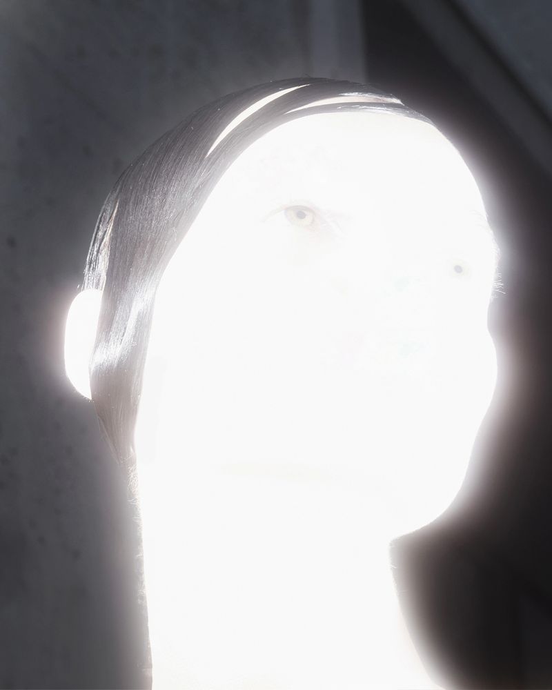 High-key portrait of an adult, fair-skinned person with short, dark hair and pale eyes, lit from the front in a grey indoor space.