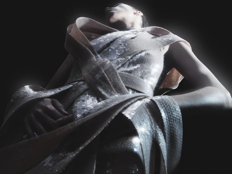 Woman in an elegant gown with shimmering detail poses in a dark studio with rim lighting highlighting the dress.