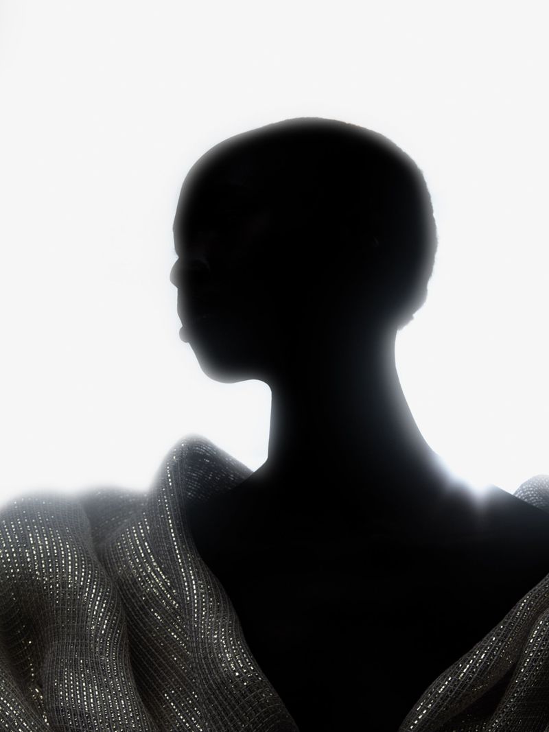 Silhouette of a bald Black woman wearing a sequined top against a stark white background in a minimalist fashion portrait.