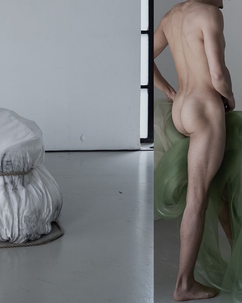 Nude adult male stands near sheer green fabric in an indoor studio setting with white walls and gray floor, and shrouded object.