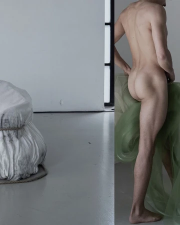 Nude adult male stands near sheer green fabric in an indoor studio setting with white walls and gray floor, and shrouded object.