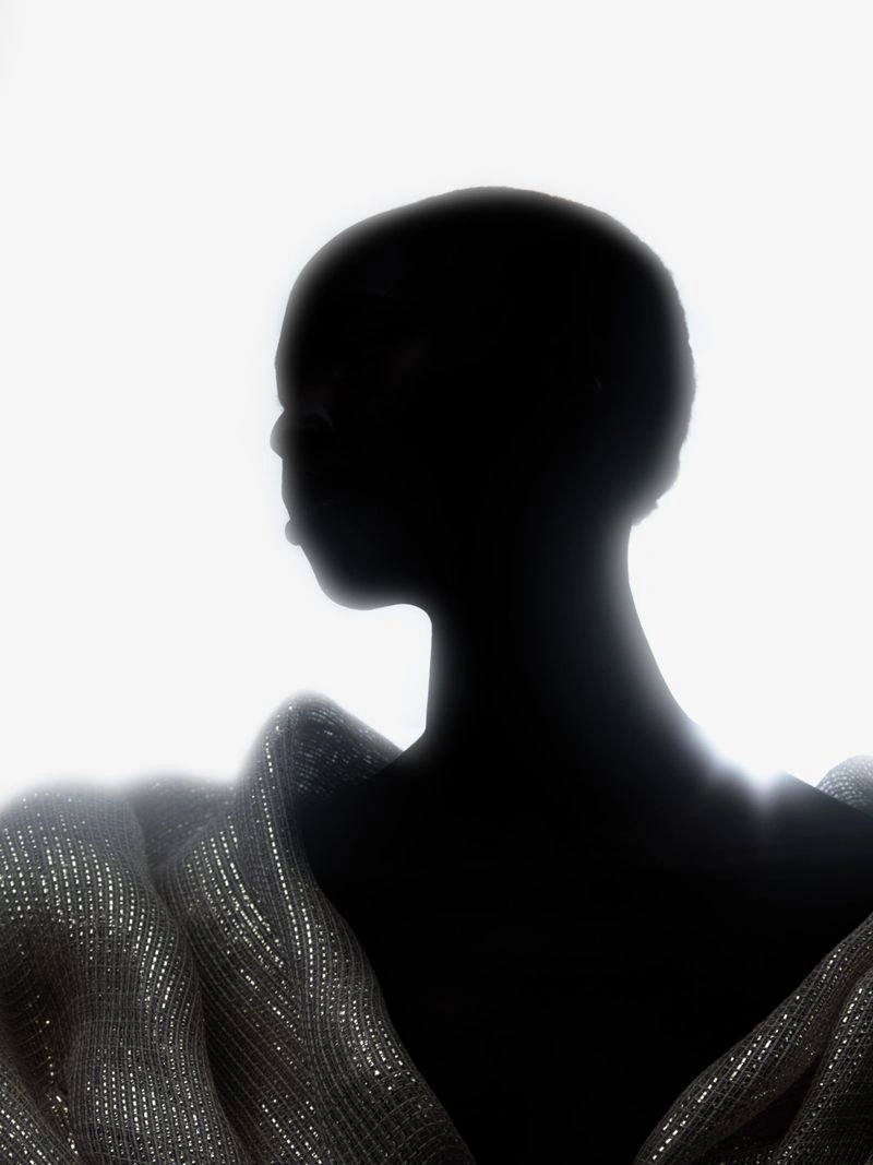 Silhouette of a model with a shaved head wearing a shimmering draped silver garment against a bright white background.