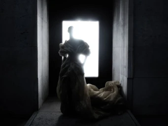 Silhouette of a model standing in a voluminous sculptural couture gown backlit by a bright rectangular doorway.