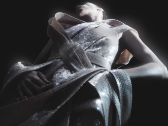 Low-angle view of a model wearing a sculptural silver sequined gown against a dark background.
