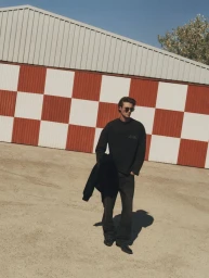 Model standing outdoors in front of a red and white checkered wall wearing a black sweatshirt and dark trousers