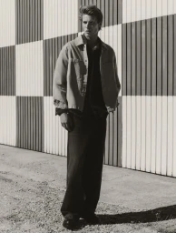 Full-length portrait of a male model wearing a light overshirt and dark trousers against a checkered paneled wall.